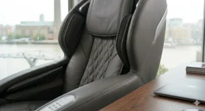 A close-up, photorealistic 4K image of the integrated armrest control panel on a grey leather office massage chair, set seamlessly into the fine-grained armrest padding within a British corporate office setting.