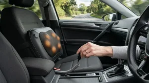 Interior of a right-hand drive car on a British country road, showing a heated massage cushion strapped to the driver's seat with its glowing nodes activated and a hand plugging it into the centre console power outlet.