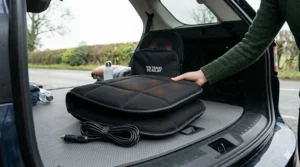 A folded heated car seat massager with its 12V power cable being placed into the boot of a car alongside travel gear.